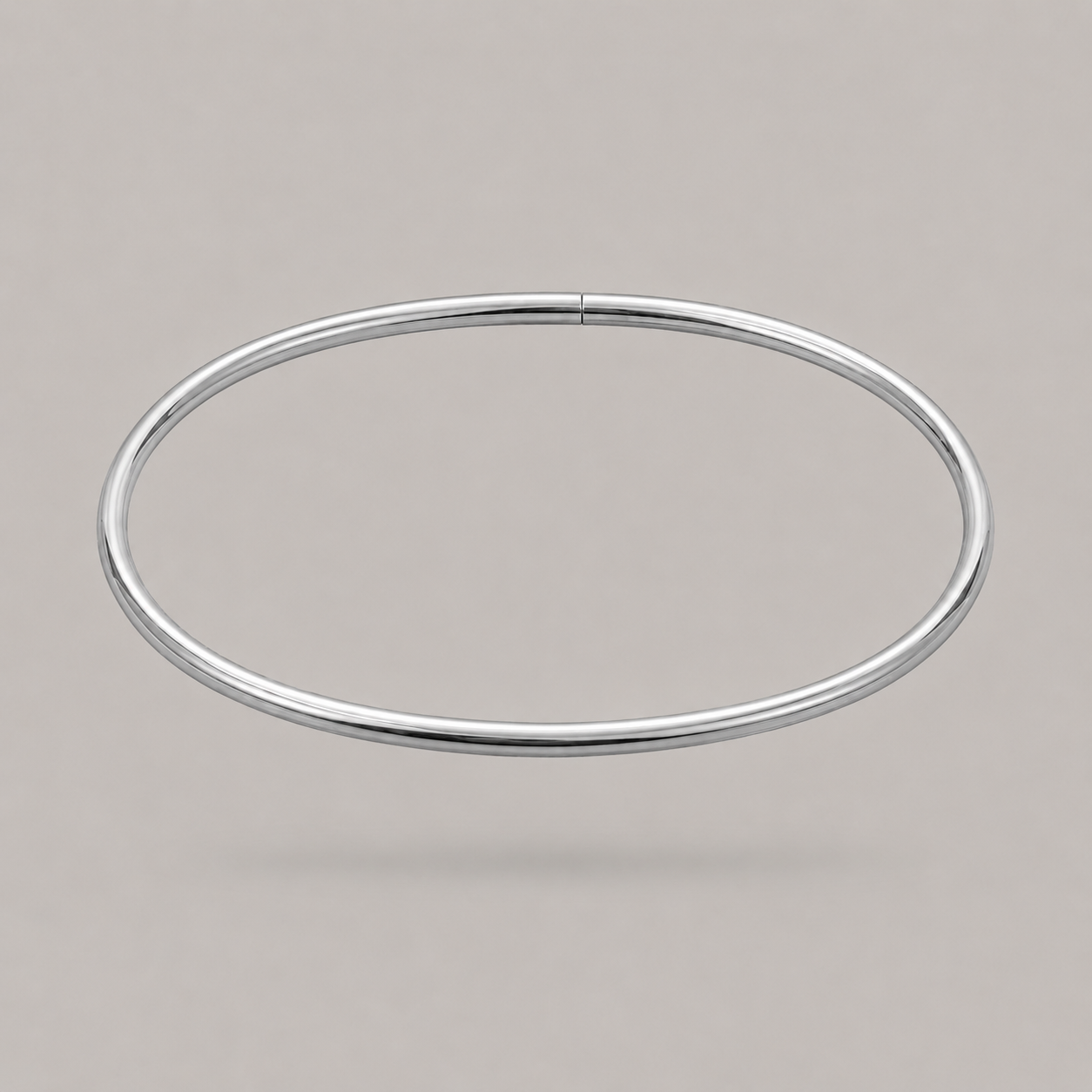 A minimalist, polished silver-toned metal bangle ankle bracelet shown against a solid, neutral warm gray background.