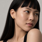 A close-up portrait of a woman with long black hair wearing a small flower gold stud earring, set against a plain light gray background.