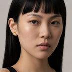 Close-up portrait of a woman with straight black hair and bangs wearing a small gold stud earring against a soft light gray background.