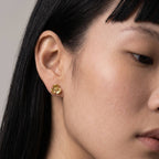 A close-up of a model's ear wearing a small gold flower-shaped stud earring against a neutral light gray background.