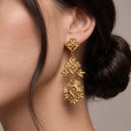 A close-up of a model's ear wearing a dangling gold earring with three textured floral clusters.
