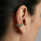 Close-up of an ear wearing a gold earring with turquoise stones against a neutral background