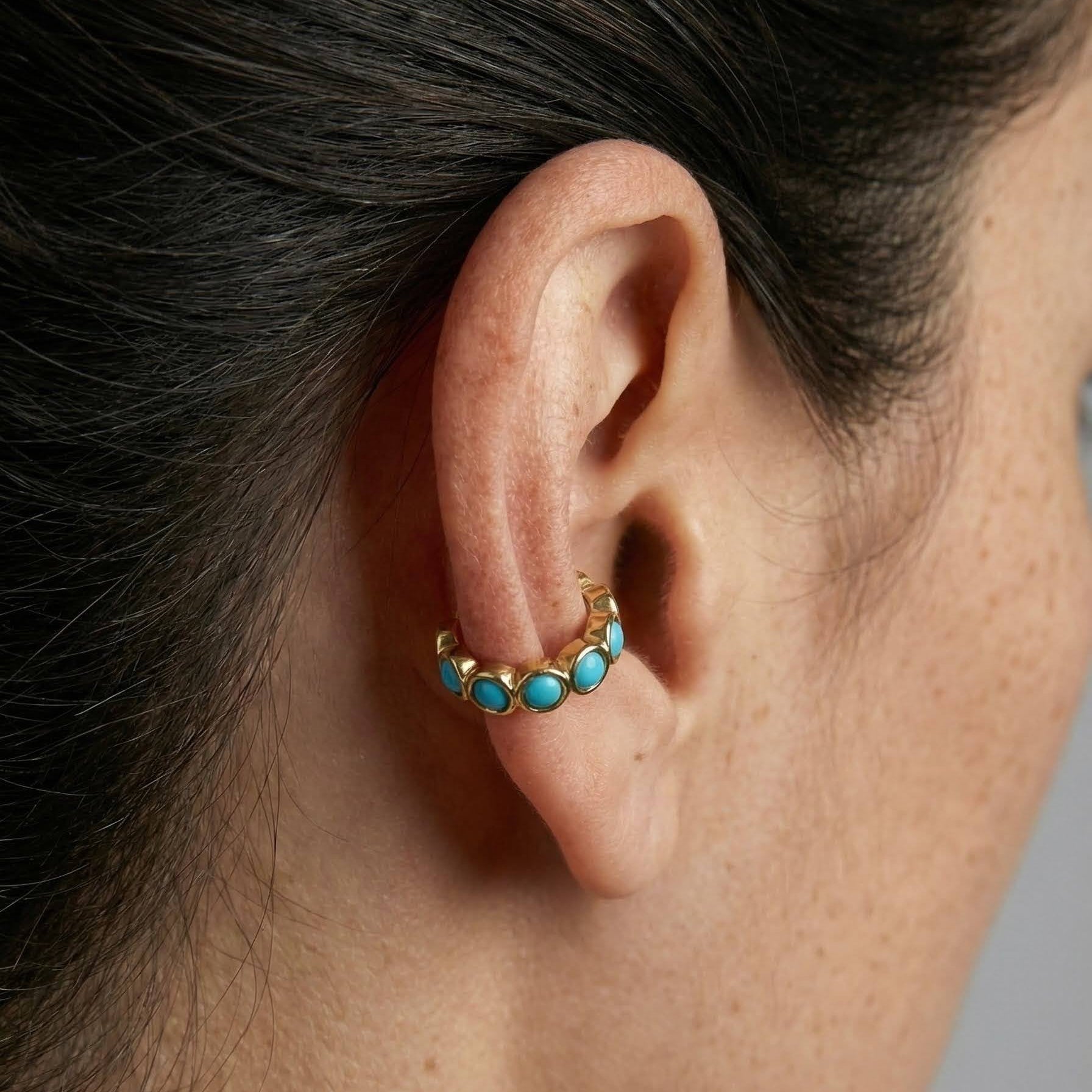 Close-up of an ear wearing a gold earring with turquoise stones against a neutral background