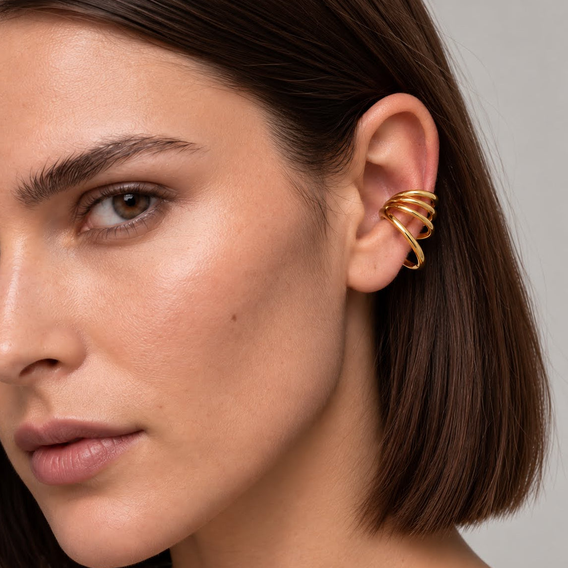 A close-up of a woman's ear wearing a gold multi-band ear cuff, set against a neutral gray background.