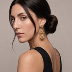 A woman with dark hair in a low bun models a pair of ornate gold drop earrings with floral details against a neutral beige background.