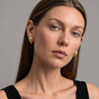 A close-up portrait of a woman wearing small gold crescent-shaped stud earrings against a plain gray background.