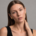 A close-up portrait of a woman wearing small gold crescent-shaped earrings against a plain gray studio background.