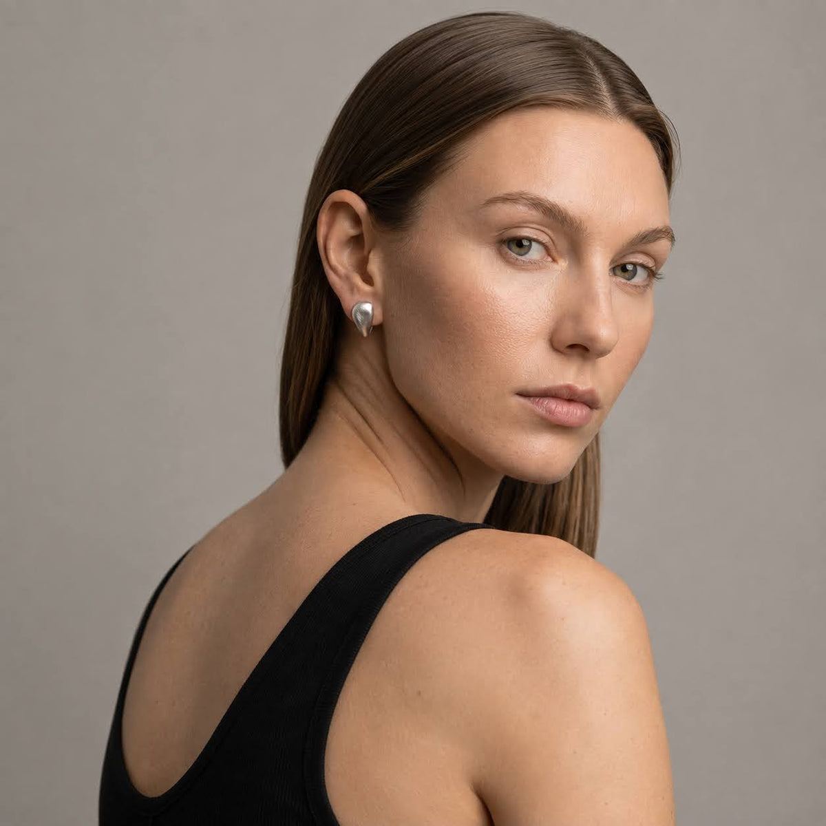 A woman with long brown hair is shown from the back and side, wearing a black sleeveless top and a small silver teardrop-shaped stud earring against a plain gray studio background.