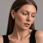 A close-up of a woman wearing small gold geometric stud earrings, shown from the chest up against a plain gray studio background.