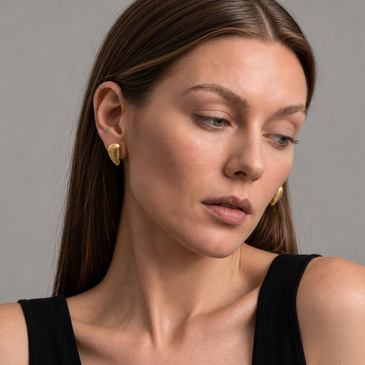 A close-up of a woman wearing small gold geometric stud earrings, shown from the chest up against a plain gray studio background.