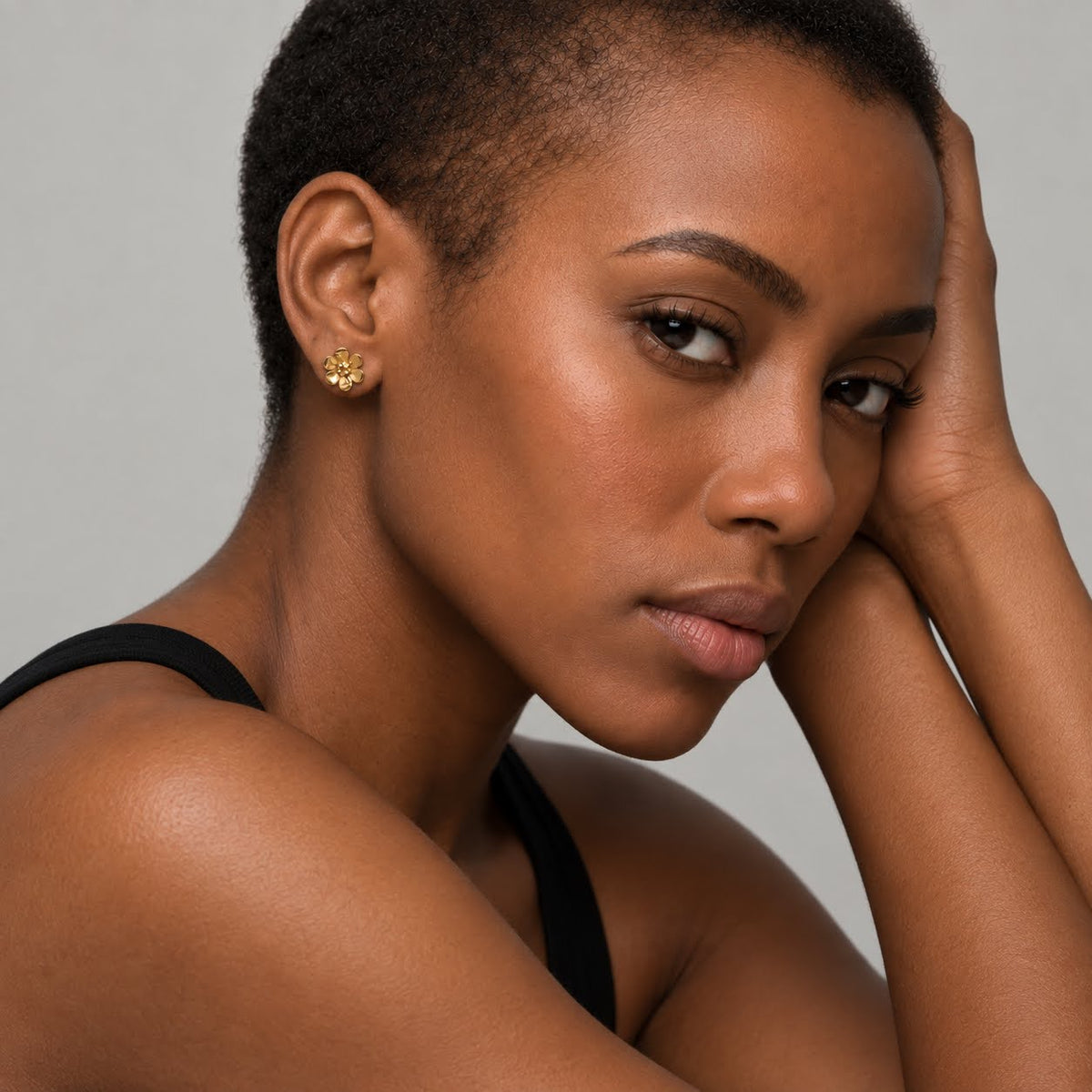 A close-up portrait of a woman with short dark hair wearing a small gold stud earring, posed against a plain light gray background.