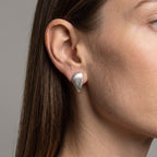 A close-up of a woman's ear wearing a textured, teardrop-shaped silver stud earring against a neutral gray background.