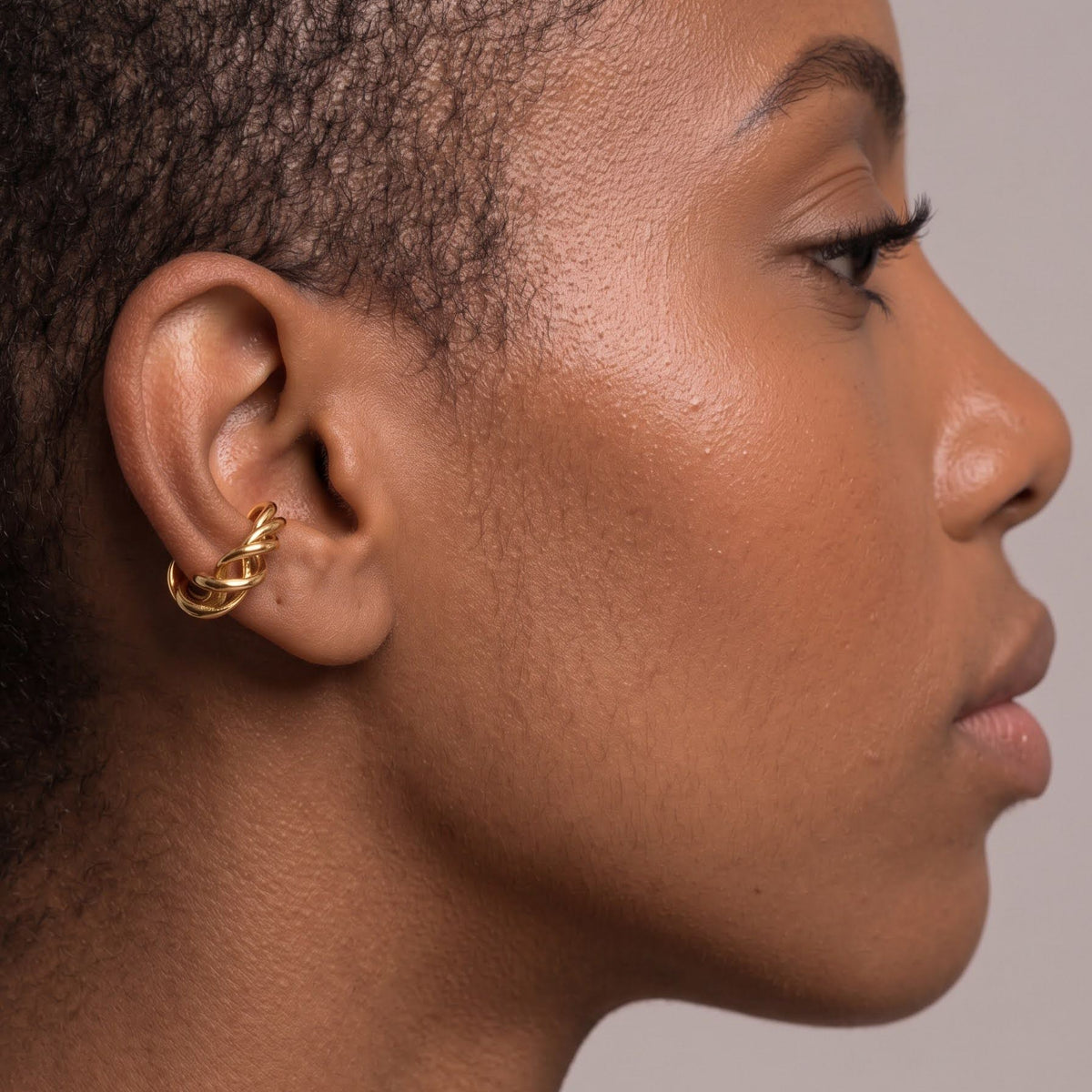 A close-up side profile of a model wearing a small gold hoop earring on her ear, set against a plain light gray background.