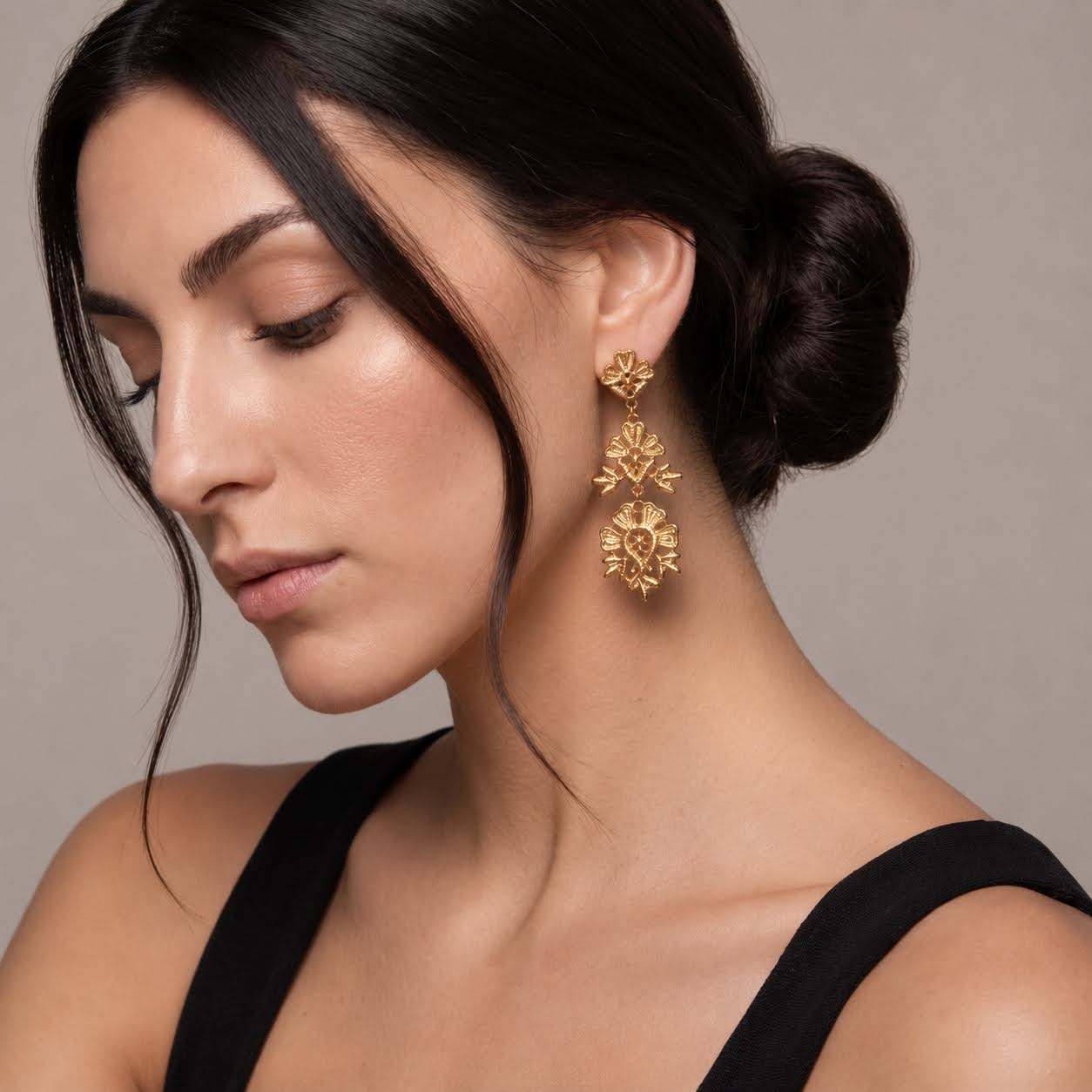 A close-up of a woman wearing a pair of gold dangle earrings with floral details, set against a neutral beige background.