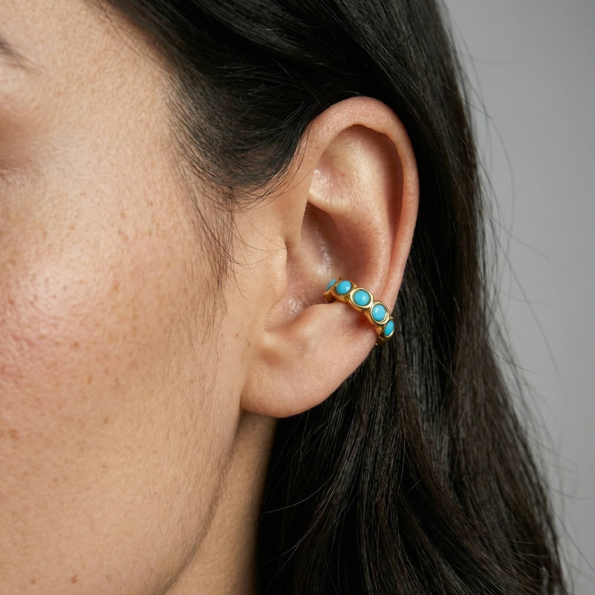 Close-up of an ear wearing a gold earring with turquoise stones against a neutral background