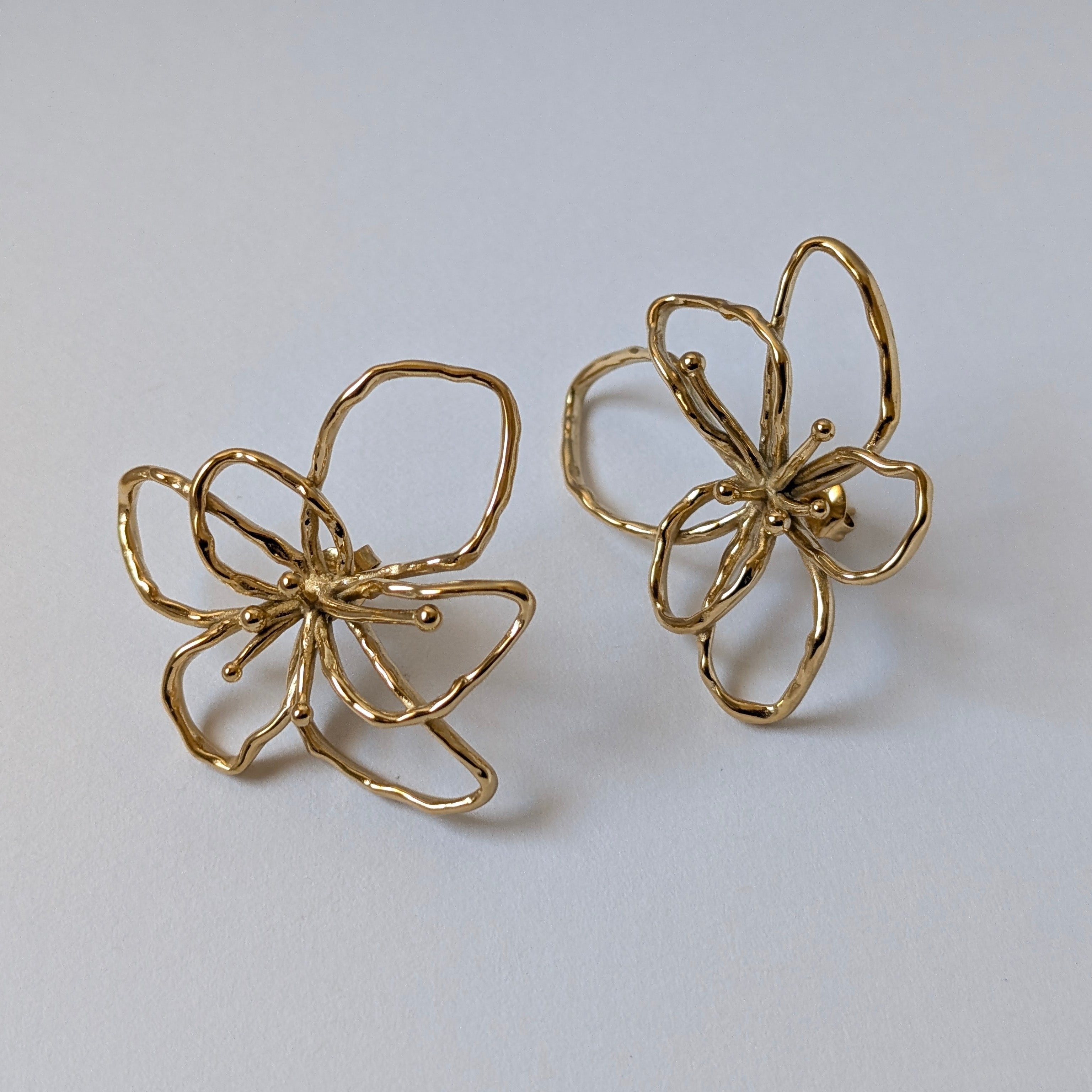 Flat‑lay of gold orchid statement earrings front side up, posts facing down on white backdrop—18k gold‑plated floral studs.