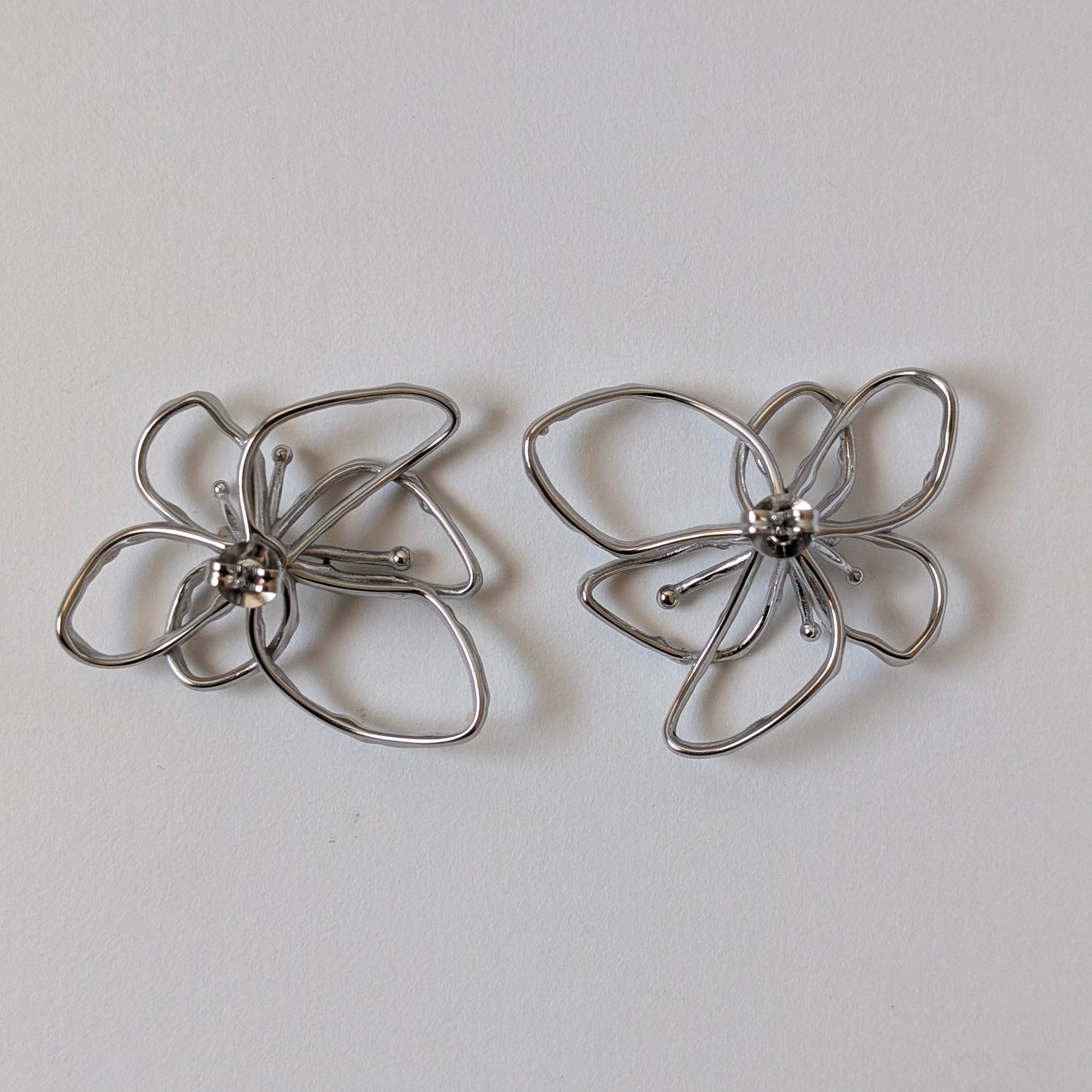 Flat‑lay of silver orchid statement earrings front side up, posts facing down on white backdrop—316 L steel petals.