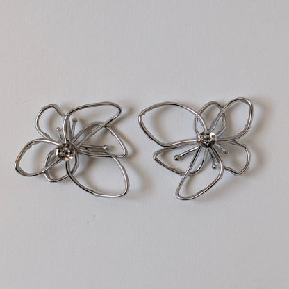 Flat‑lay of silver orchid statement earrings front side up, posts facing down on white backdrop—316 L steel petals.